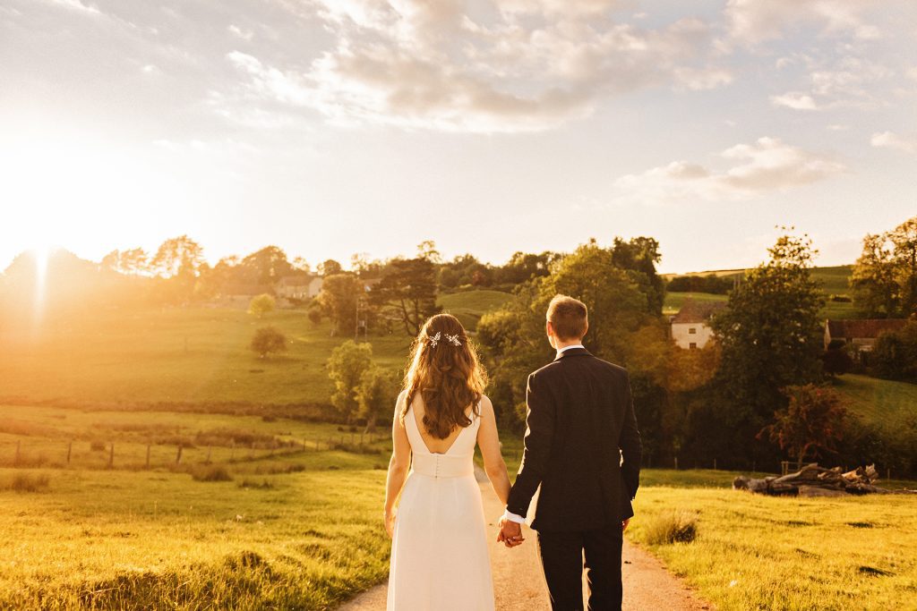 Rebecca & Rhydian Bride and groom with their backs to the camera, holding hands looking back on Kingscote Barn, Photo by Gloucestershire wedding photographer Blooming Photography.